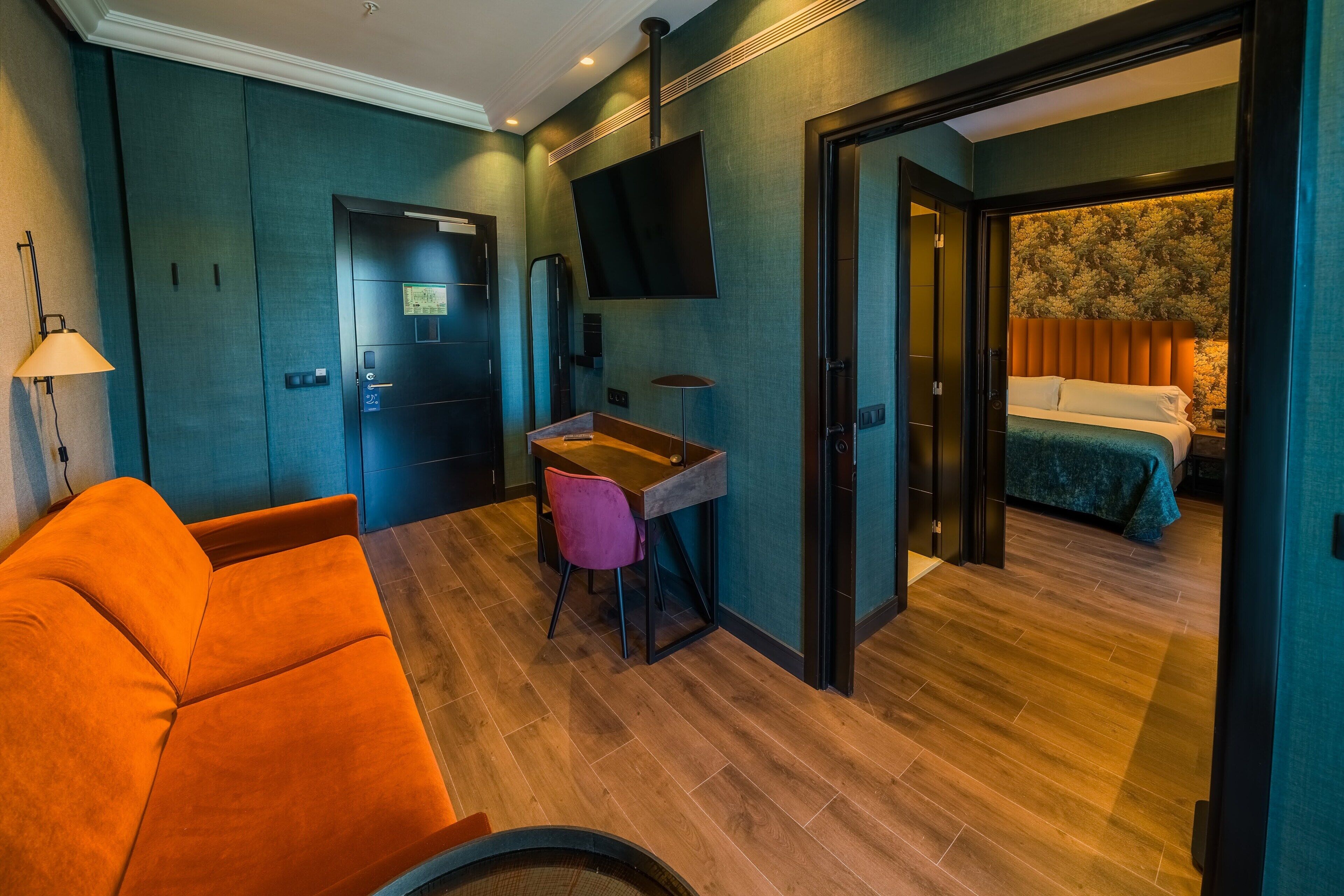 A modern hotel room with a sofa, a desk, a chair, a television, and a bed with a floral headboard.