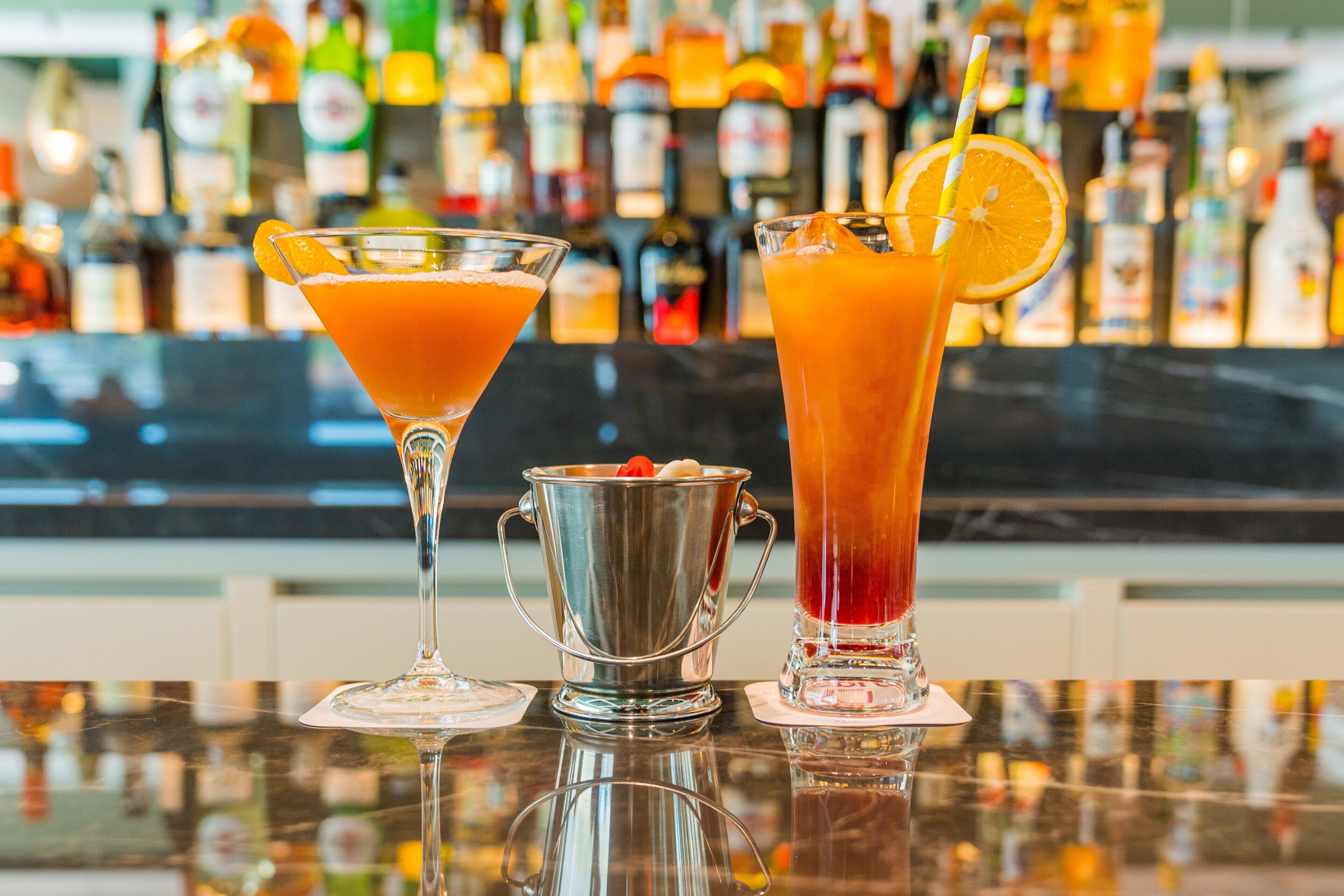 Two cocktails on a bar counter with a background of various liquor bottles.