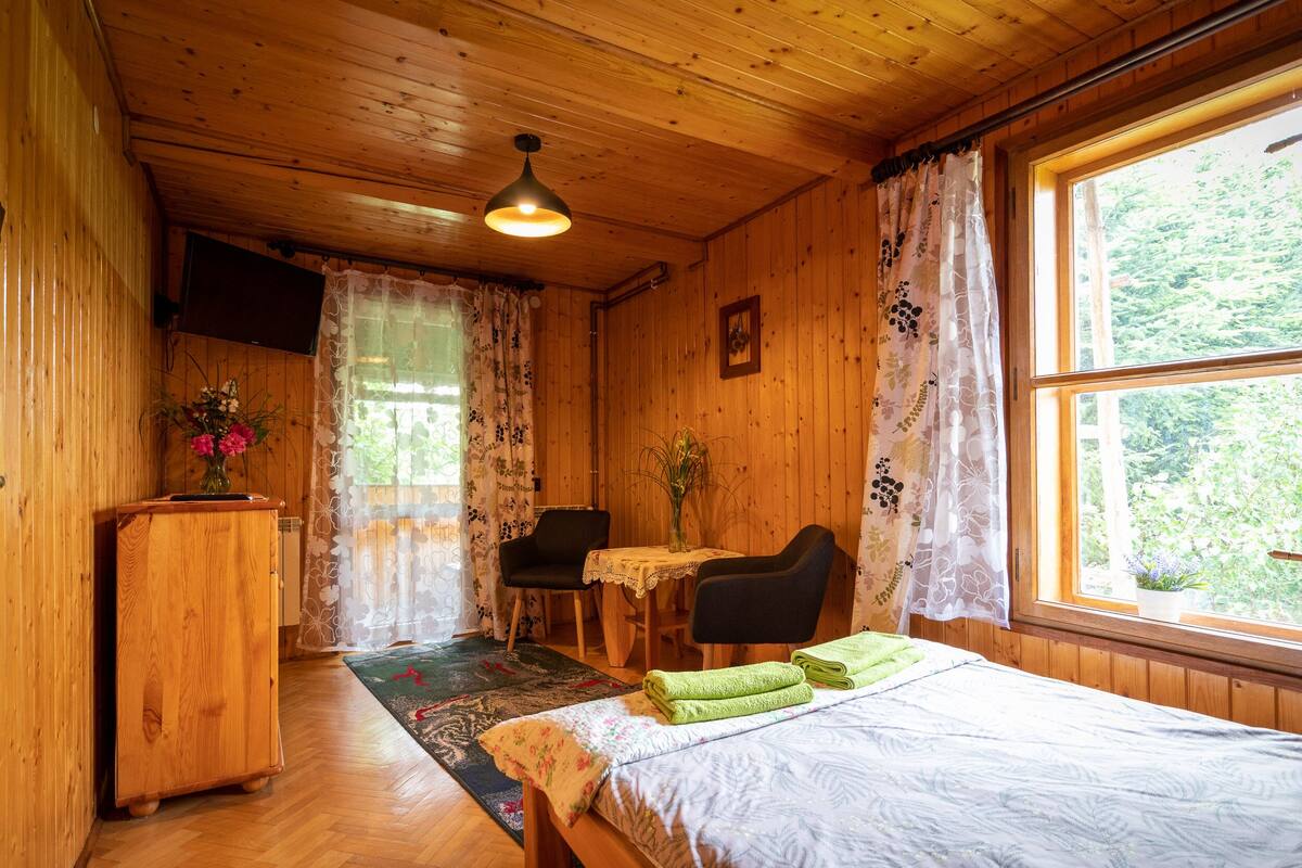 Habitación de cabaña de madera con cama, escritorio, silla, televisión y ventana con vista a follaje.