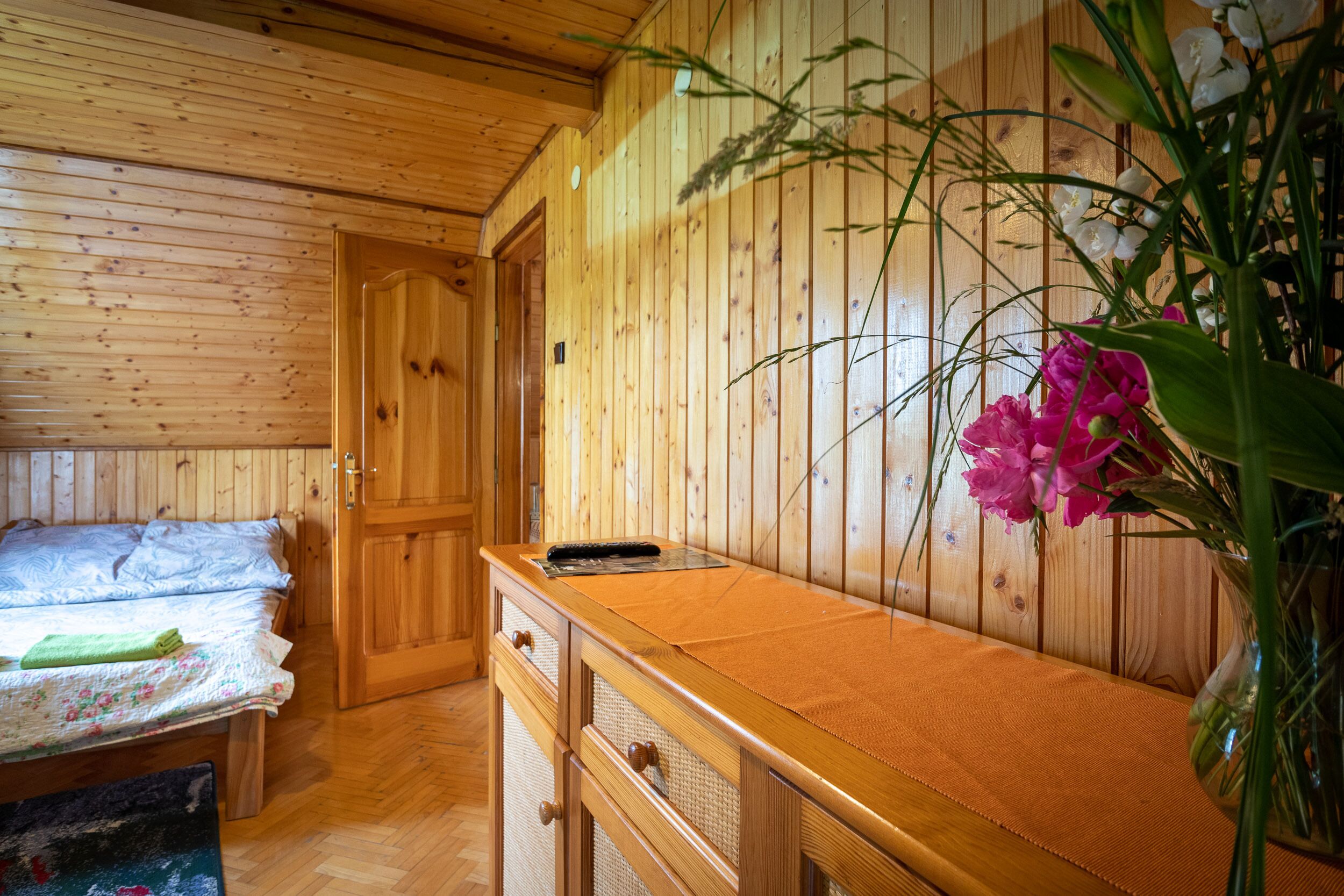 A wooden cabin interior with a bed, a wooden dresser, and a vase with flowers.
