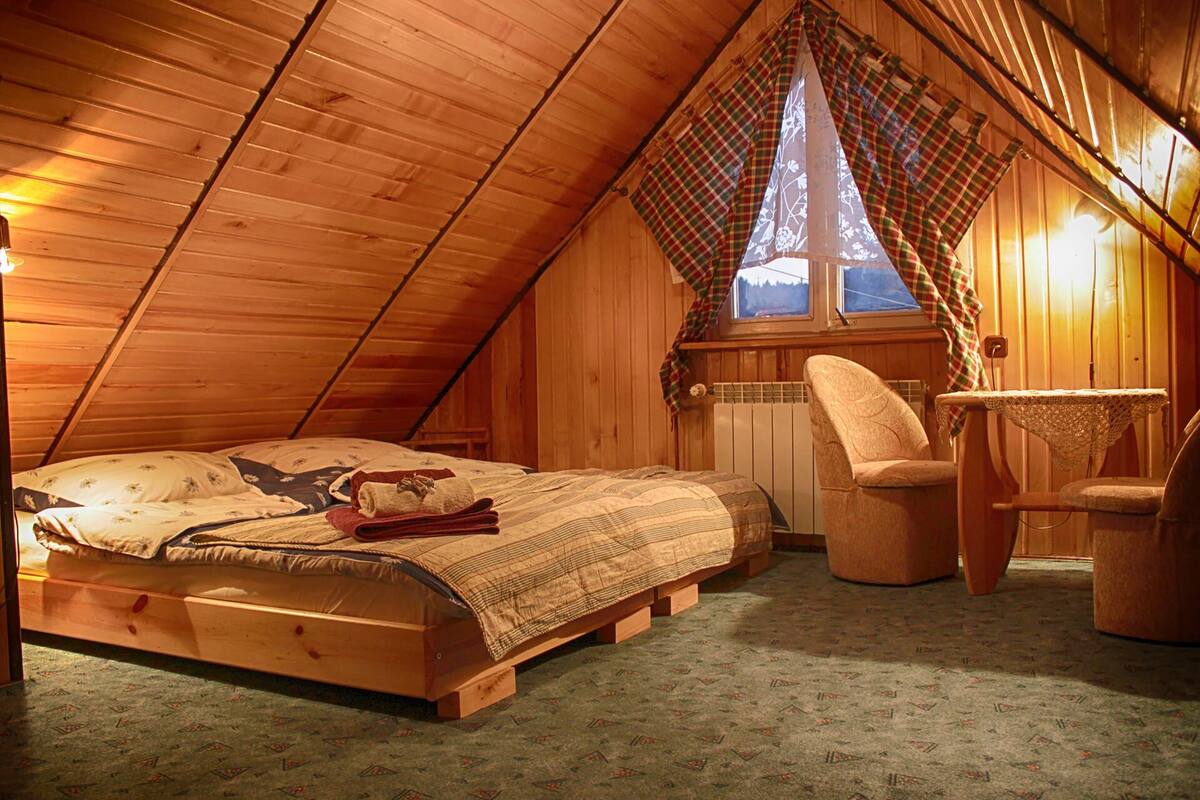 Un dormitorio cálido en el ático con techo inclinado de madera, una cama con ropa de cama estampada, una silla y una mesita.