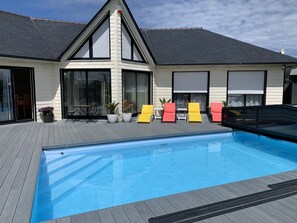 Piscina com deck de madeira, área de varanda coberta e uma casa com janelas amplas e telhado triangular.