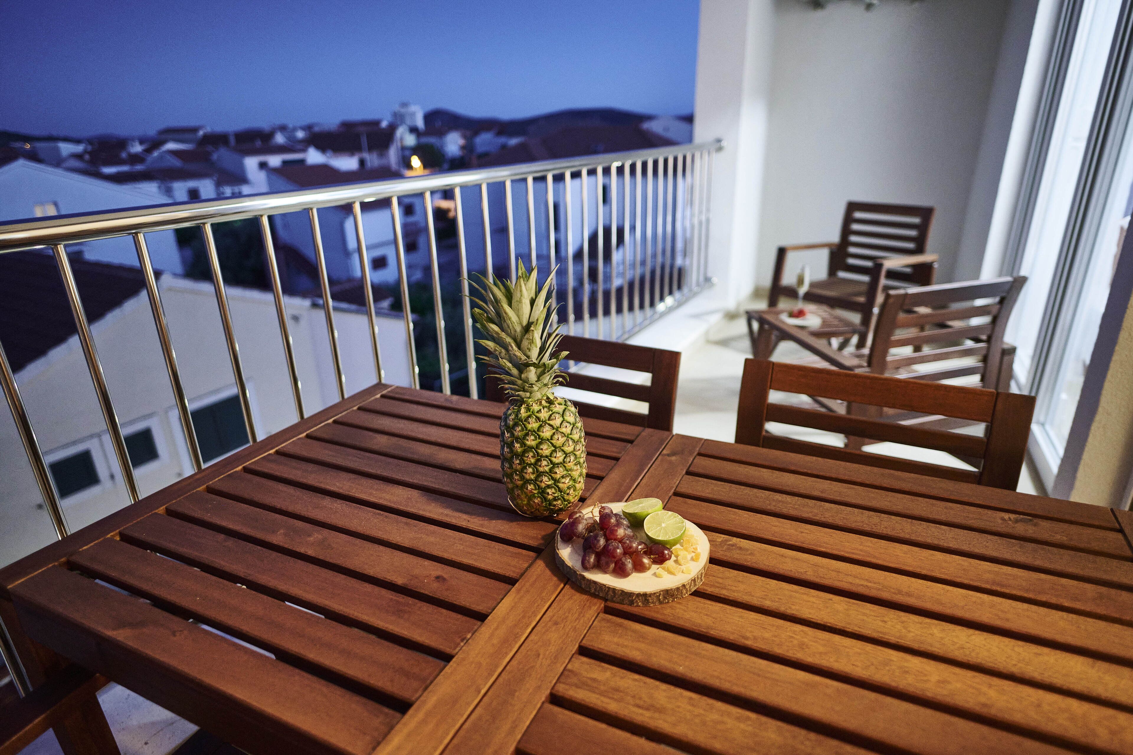 A wooden table with a pineapple and a plate of fruit on a balcony overlooking a cityscape.