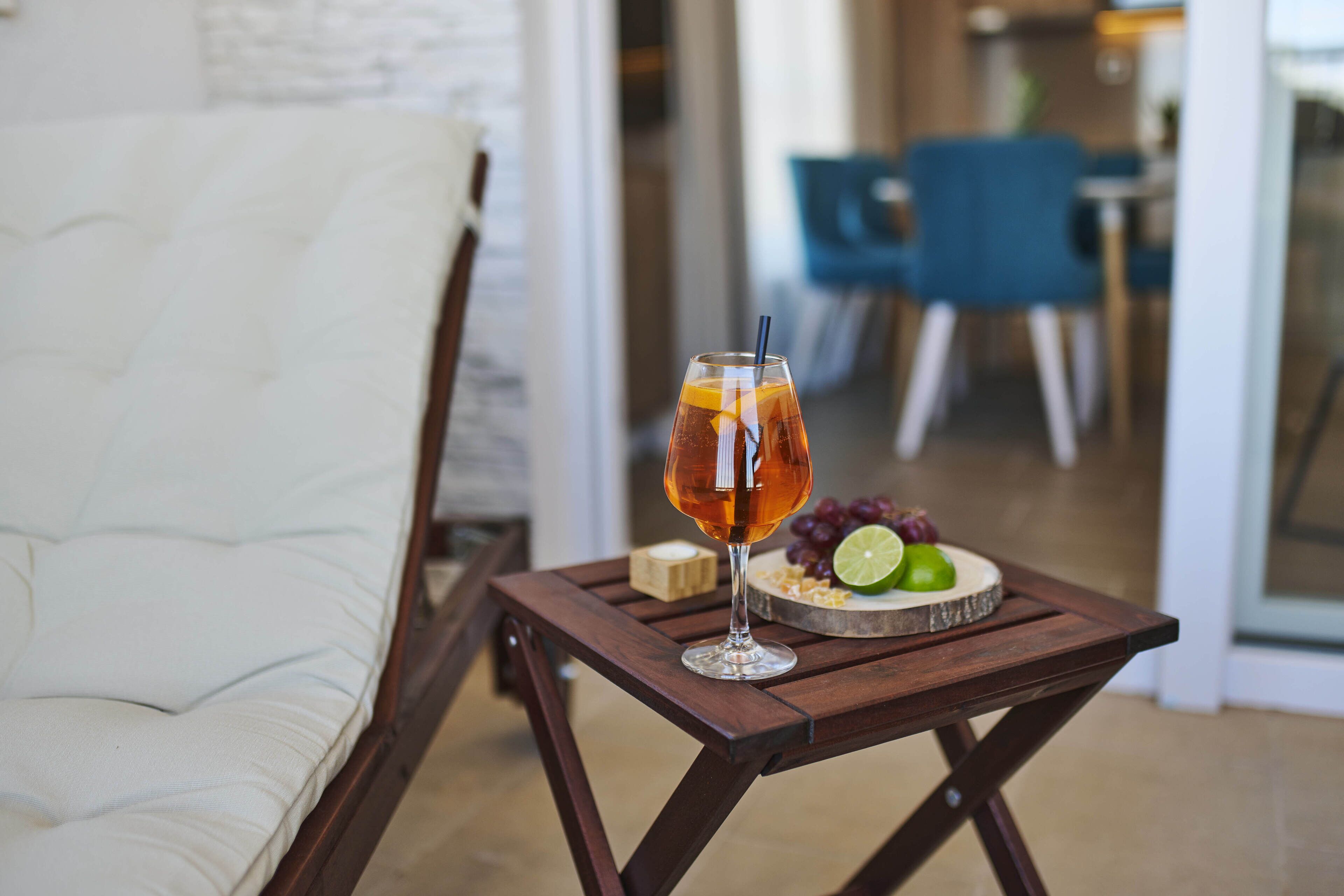 A glass of amber-colored beverage on a wooden tray with a lime and grapes, placed on a small table in a room with a white couch and blue chairs.
