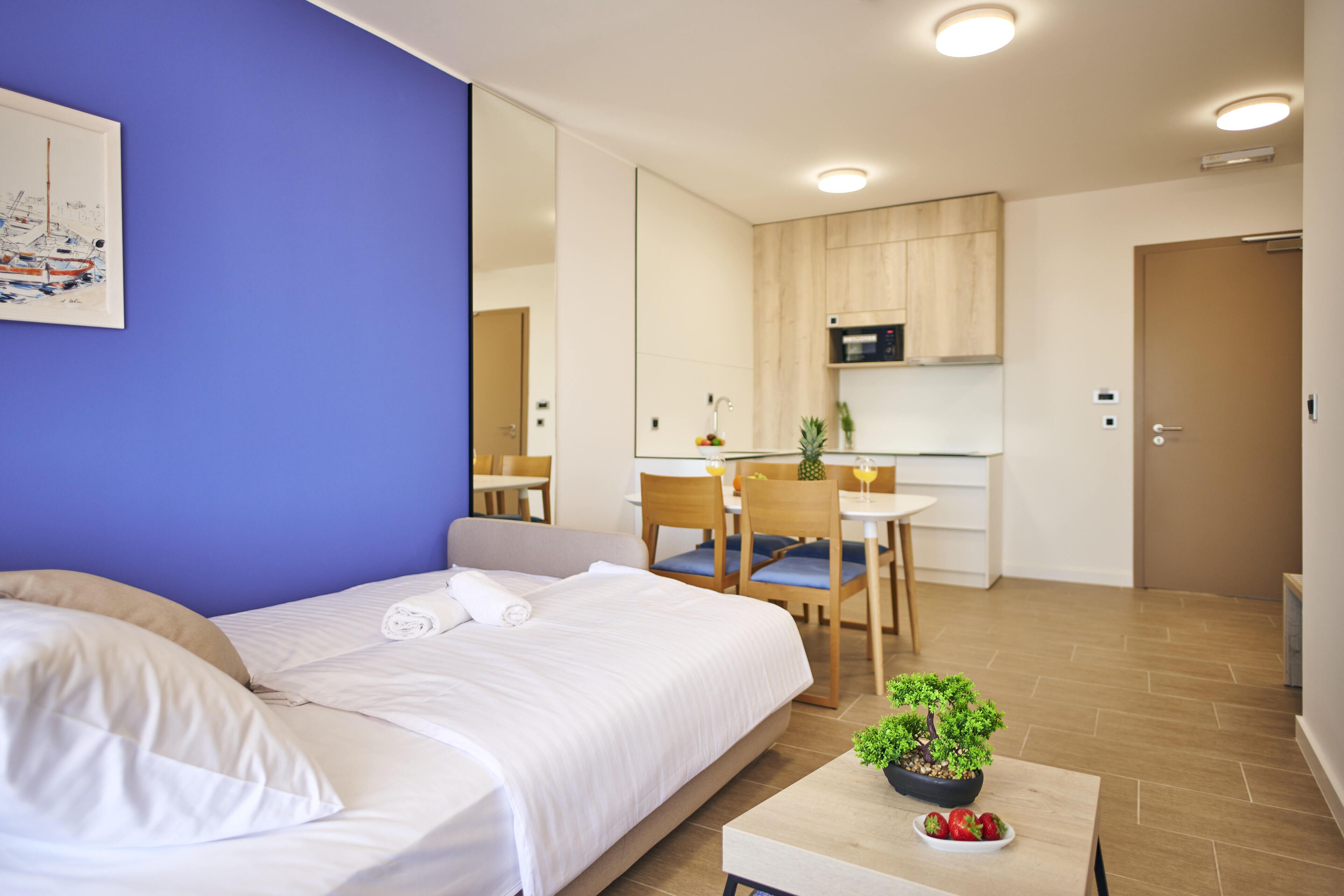 A hotel room with a bed, a small table with a plant, and a kitchenette in the background.