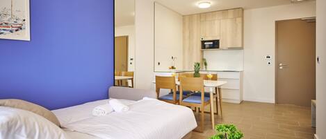 A hotel room with a bed, a small table with a plant, and a kitchenette in the background.