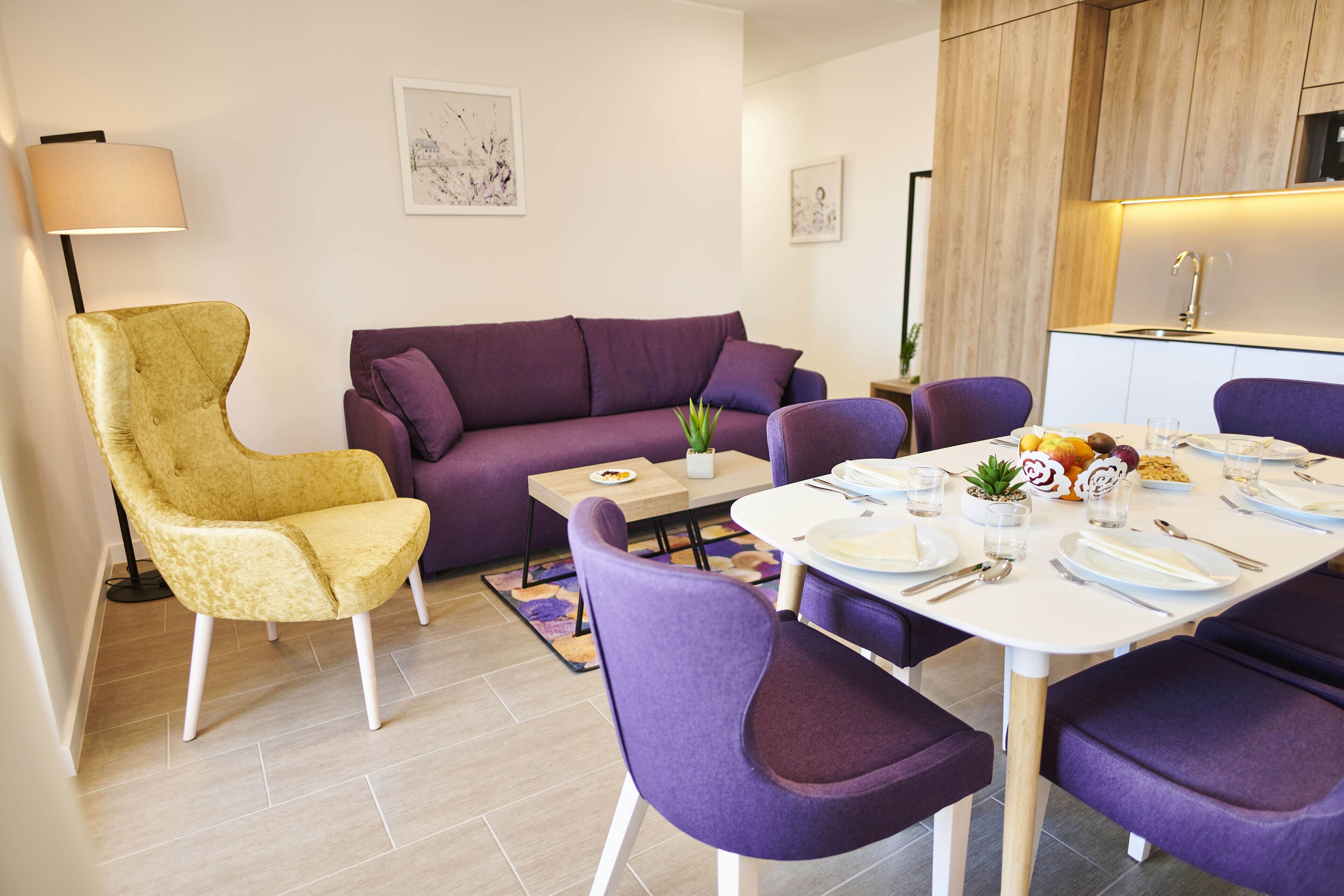 Un salon moderne avec un canapé, un fauteuil jaune, une table à manger dressée pour quatre personnes et, au fond, un coin cuisine.