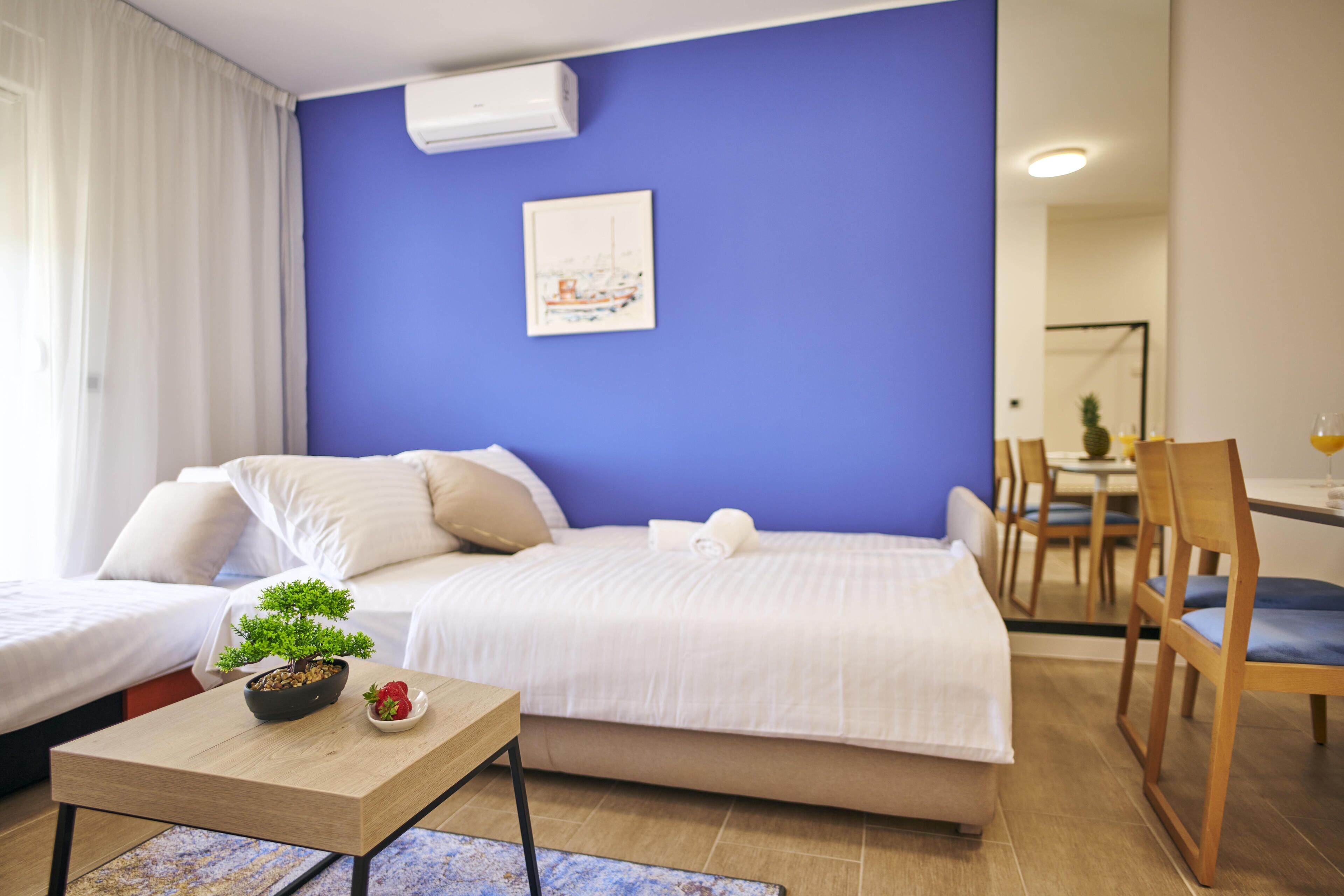 Hotellihuone, jossa on sänky, pieni pöytä, jonka päällä on kasvi ja hedelmiä, seinällä taulu sekä ruokailutila, jossa on tuoleja ja pöytä.