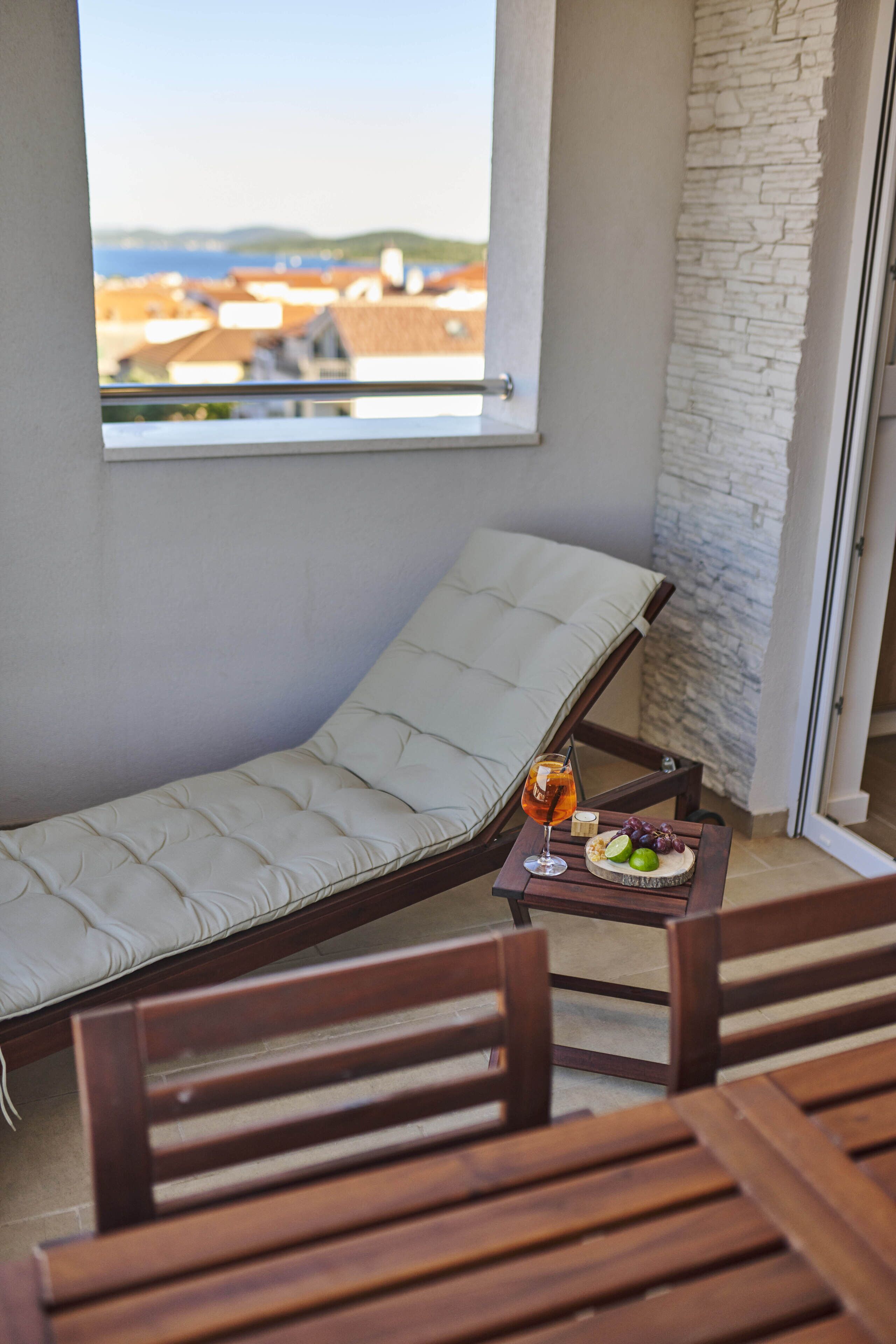 Un balcon avec un fauteuil confortable, une table avec une boisson et des fruits, et une vue sur les bâtiments et les collines.