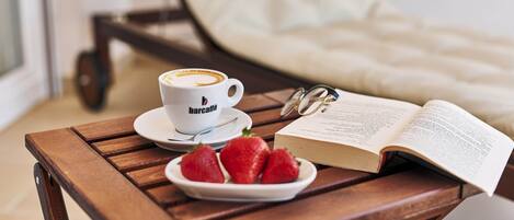 A wooden table with a cup of coffee, a plate of strawberries, a book, and a pair of glasses.