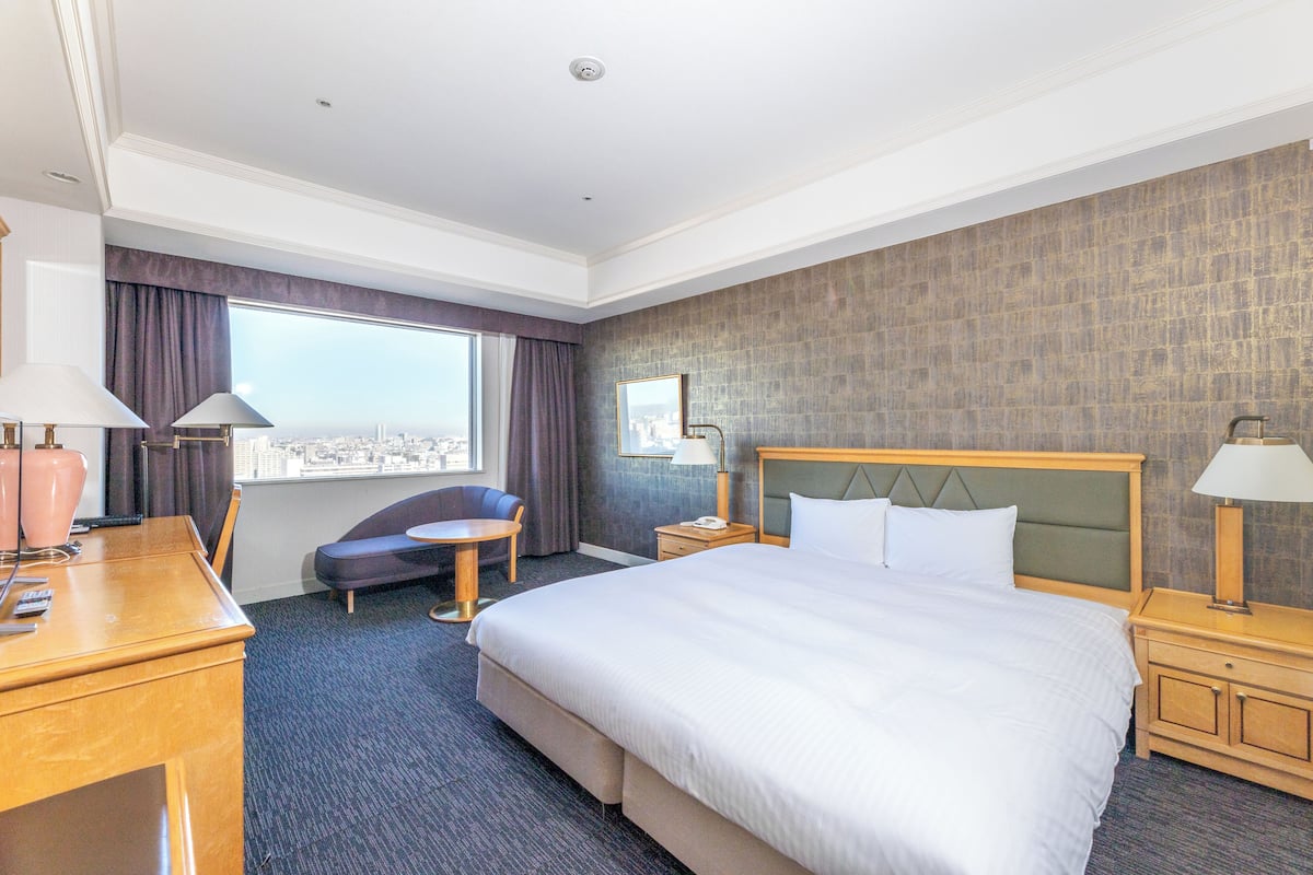 Une chambre d’hôtel avec un grand lit, un bureau, une chaise, une petite table et une fenêtre avec des rideaux.