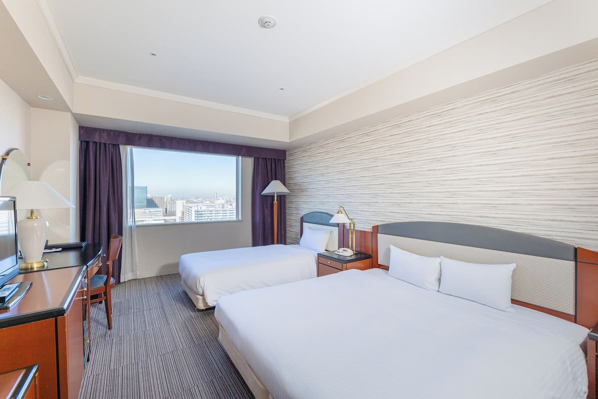Une chambre d’hôtel avec deux lits, un bureau, une chaise, une lampe et une fenêtre donnant sur la ville.