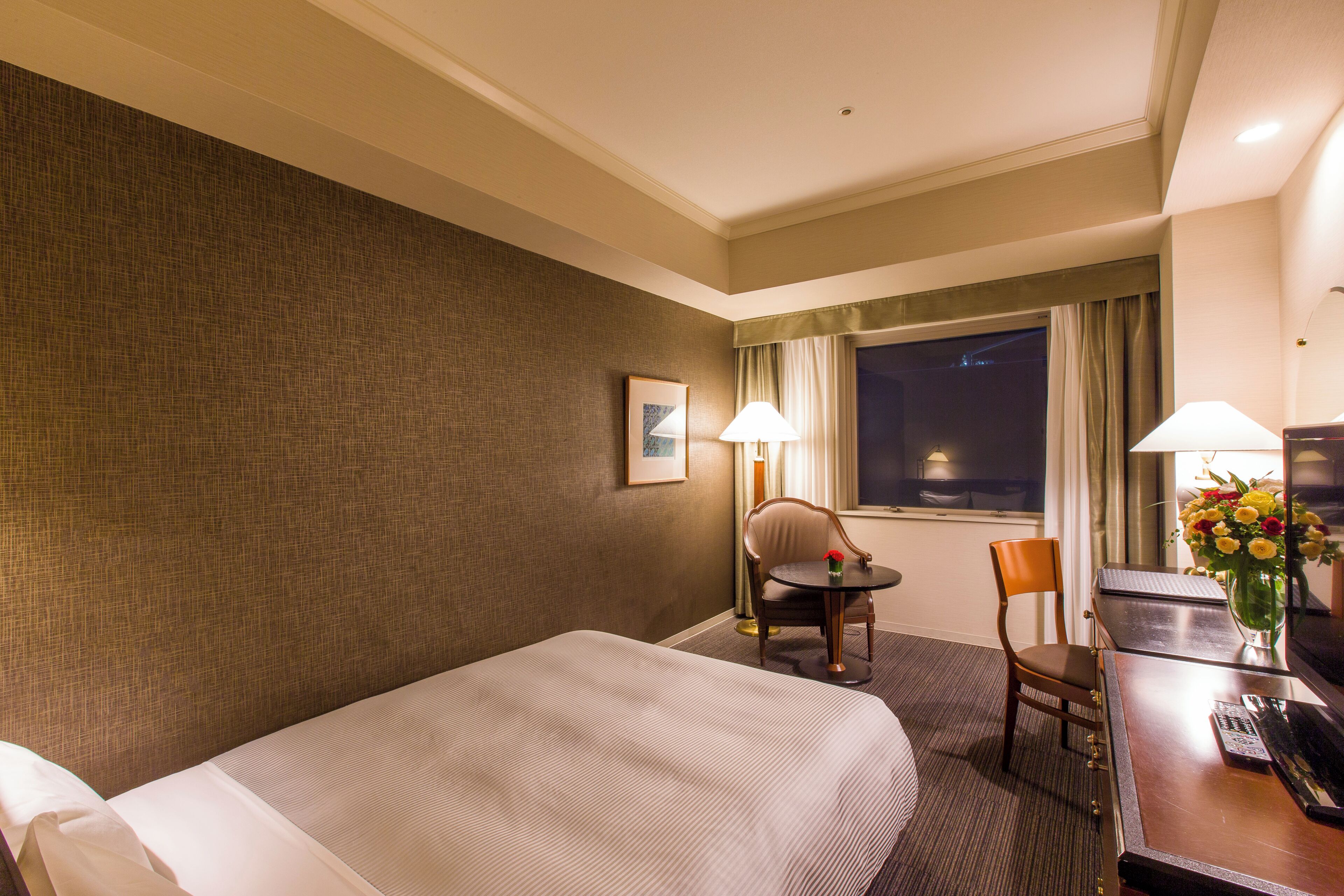 Habitación de hotel con una cama, dos sillas, una mesita, un escritorio, una lámpara, un jarrón con flores y una ventana con cortinas.