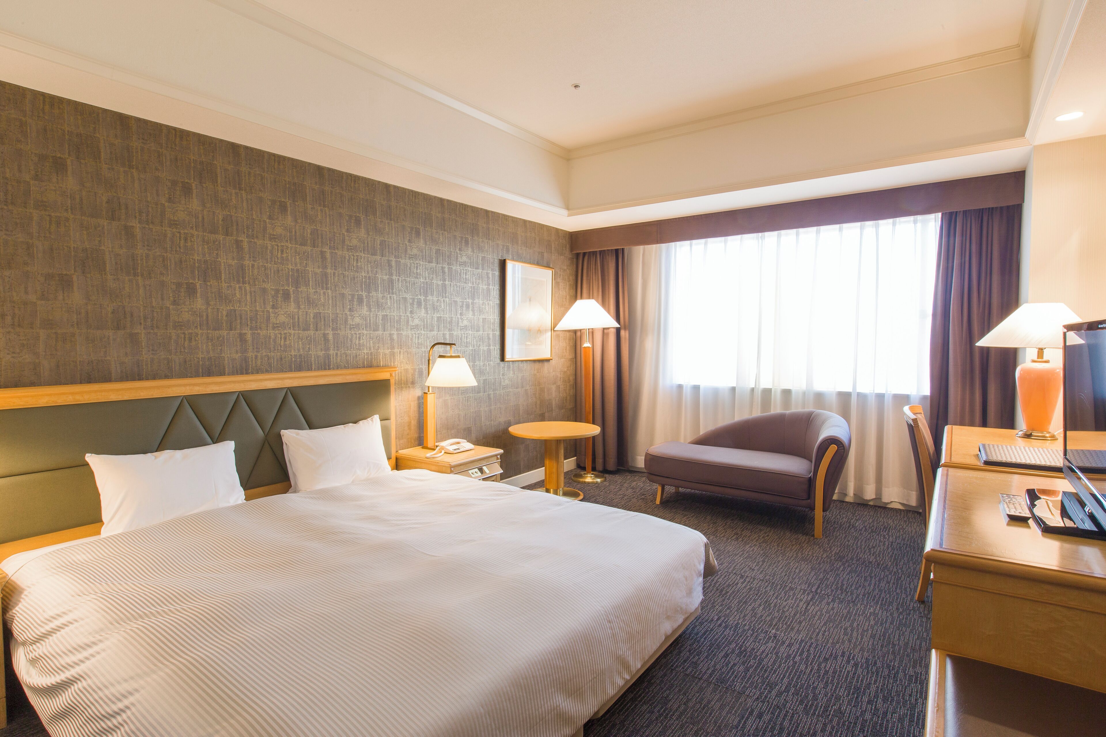 A hotel room with a large bed, a desk with a computer, a chair, a small round table, and a window with curtains.