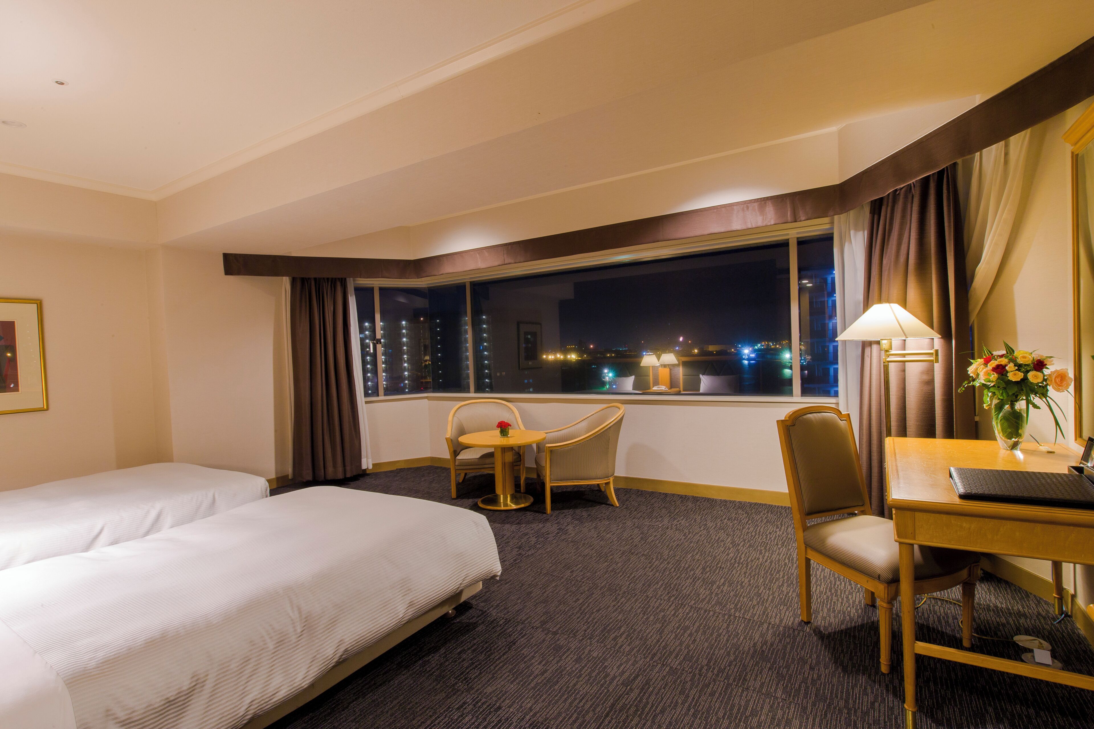 A hotel room with two beds, a desk with a lamp, a chair, and a window with a city view.