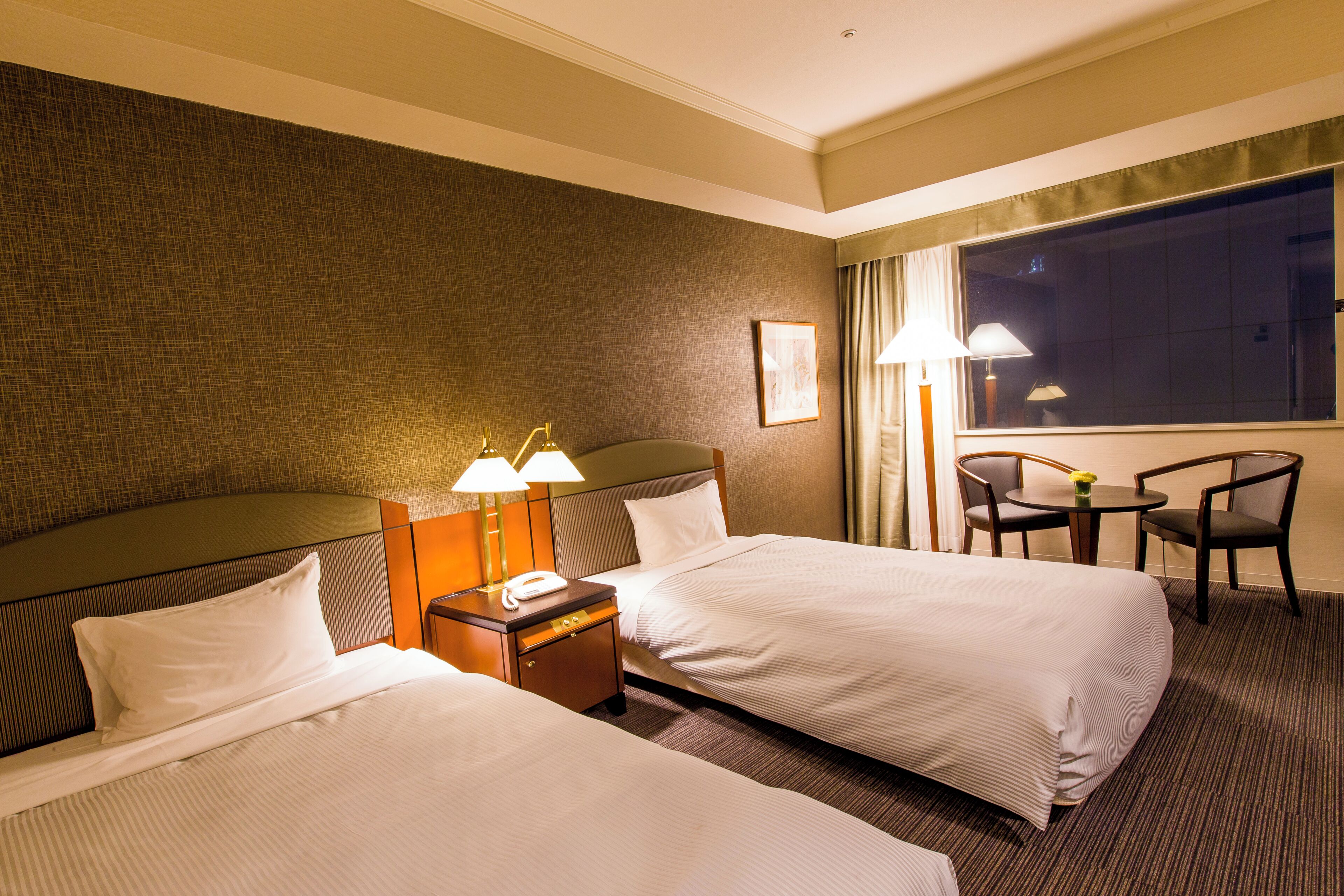 Quarto de hotel com duas camas, mesa de cabeceira com luminárias, pequena mesa com cadeiras e uma janela grande.