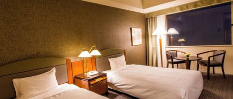 Quarto de hotel com duas camas, mesa de cabeceira com luminárias, pequena mesa com cadeiras e uma janela grande.
