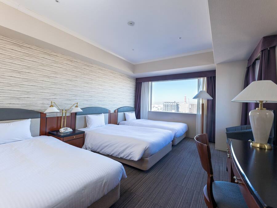 Habitación de hotel con tres camas, un escritorio, una silla, una lámpara y vista a la ciudad.