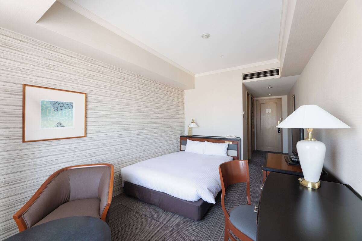 Une chambre d’hôtel avec un lit, deux chaises, un bureau, une lampe et un tableau encadré au mur.