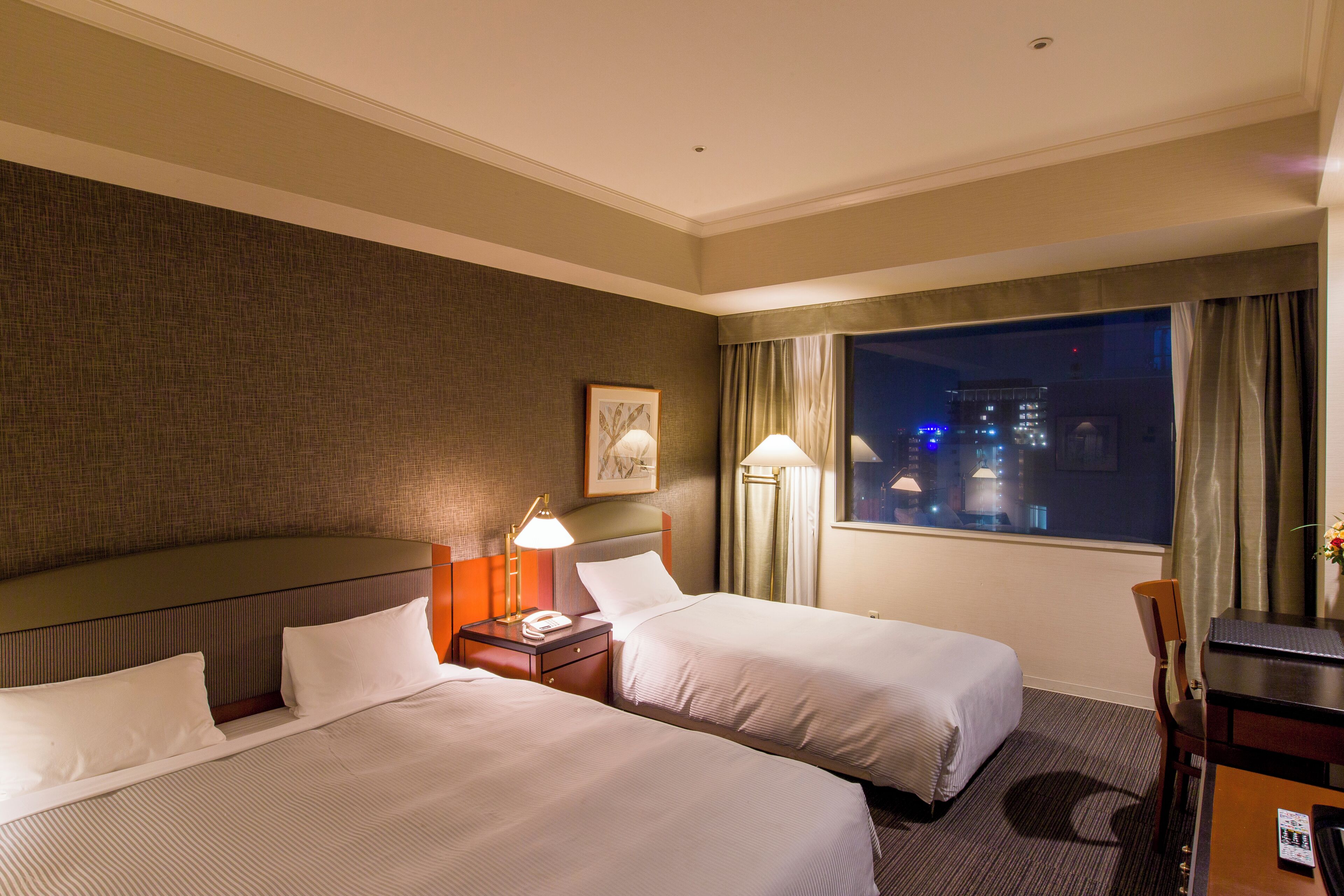 A hotel room with two beds, a desk, a chair, a nightstand with a lamp, and a window with a view of the city at night.