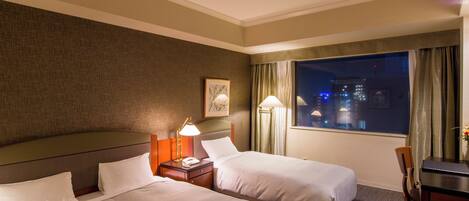 A hotel room with two beds, a desk, a chair, a nightstand with a lamp, and a window with a view of the city at night.