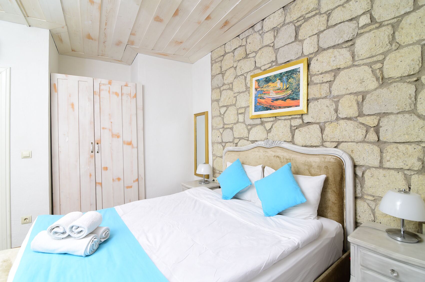 A bedroom with a stone wall, a bed with blue pillows, a nightstand, and a painting above the bed.