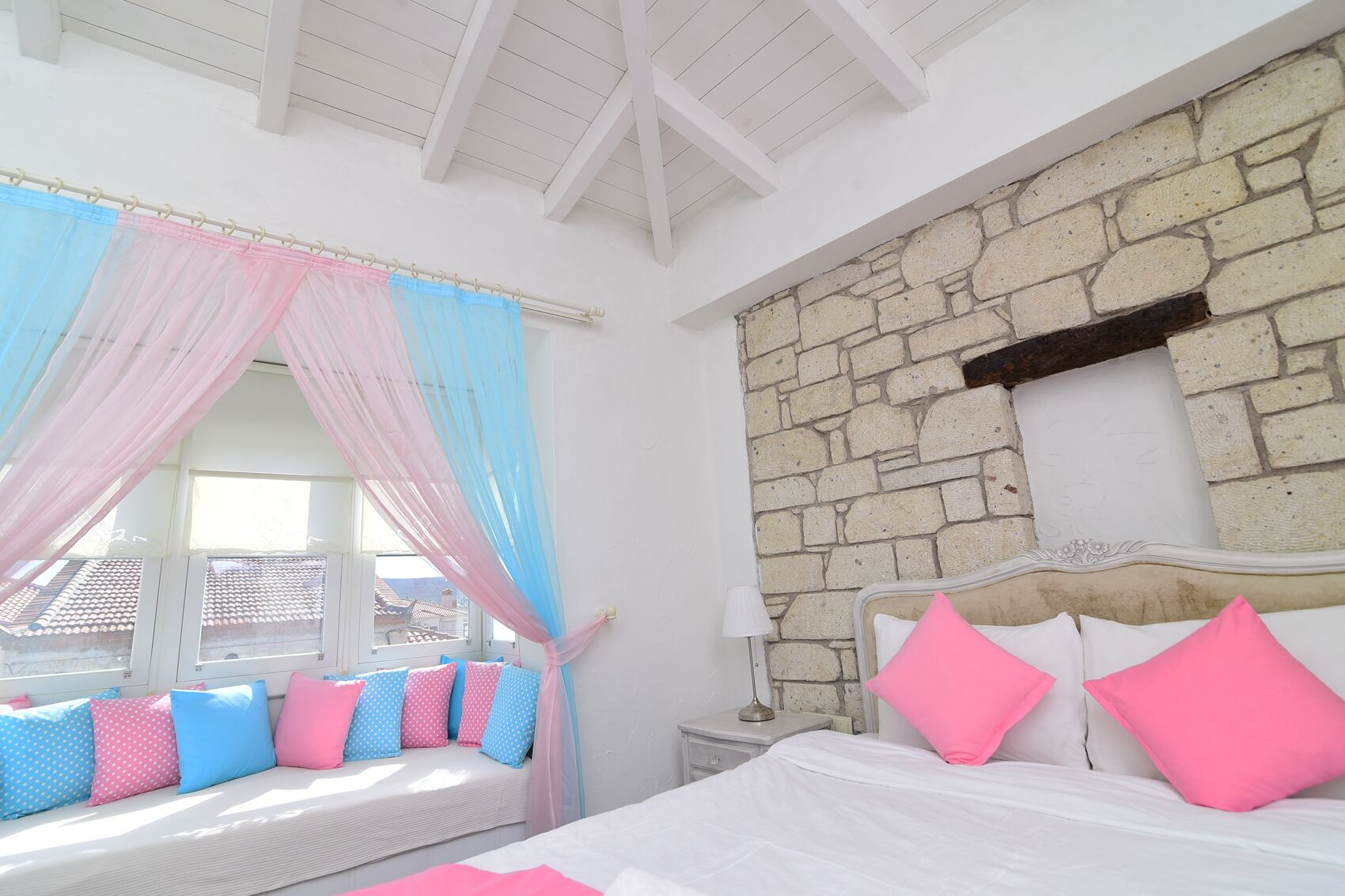 Un dormitorio con pared de piedra, cama con ropa de cama rosa y blanca, ventana con vistas y una banca con cojines de colores.