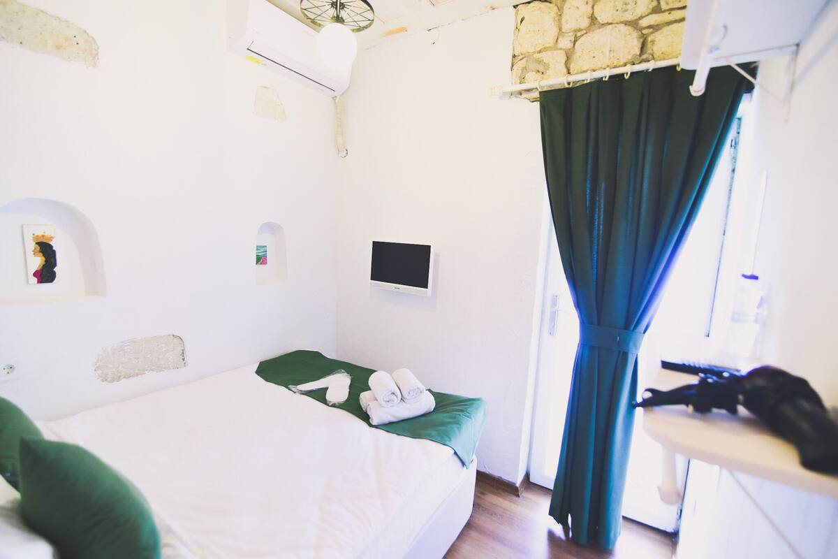 A bedroom with a bed, a small table, a chair, a window with curtains, and a wall-mounted TV.