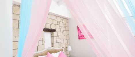 A bedroom with a bed, a nightstand, a stone wall, and a window with sheer curtains.