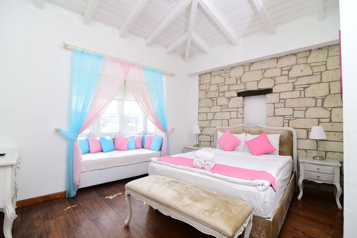 A bedroom with a stone wall, a bed with pink and white bedding, a bench, and a window with blue and pink curtains.