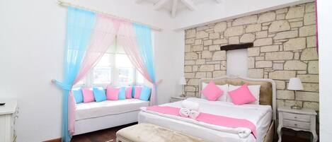 A bedroom with a stone wall, a bed with pink and white bedding, a bench, and a window with blue and pink curtains.