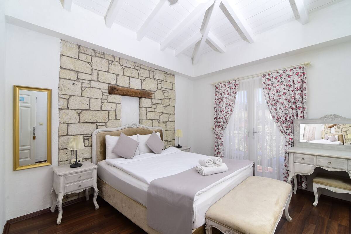 A bedroom with a stone wall, a bed with white bedding, a nightstand with a lamp, a dresser, and a bench.