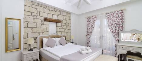 Un dormitorio con pared de piedra, una cama con ropa blanca, una mesita de noche con lámpara, un aparador y un banco.