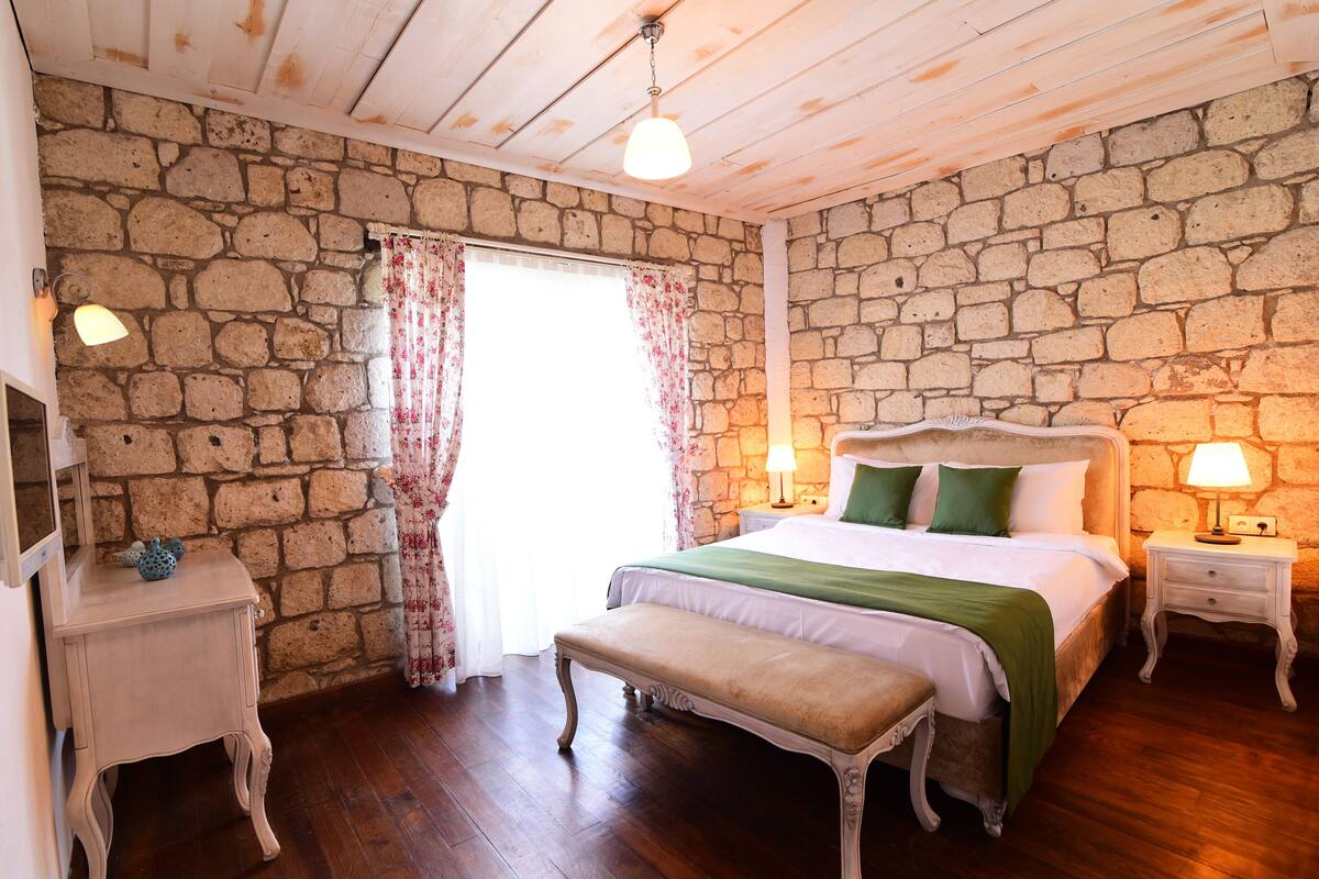 A bedroom with a stone wall, wooden floor, a bed with green bedding, a bench, a nightstand with a lamp, and a window with floral curtains.