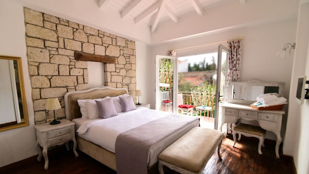 A bedroom with a stone wall, a bed with a bench, a desk, and a balcony with a view. - Zübeyde Hanım Alaçatı (Cesme)