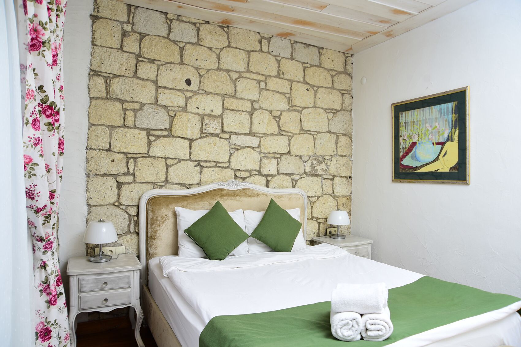 Un dormitorio con pared de piedra, cama con almohadas verdes, un cuadro en la pared y cortina estampada.