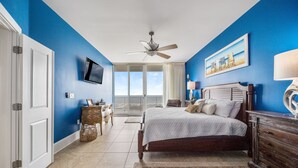 A bedroom with a large bed, a ceiling fan, a TV, and a view of the sky through the window.
