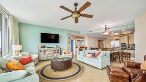 A spacious living room with a ceiling fan, a television, and a dining area with a kitchen in the background.