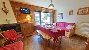 A cozy dining area with a wooden table set for four, a red sofa, and a television mounted on the wall. - Cozy 2-room apartment, 6 beds, near slopes (Le Grand-Bornand)