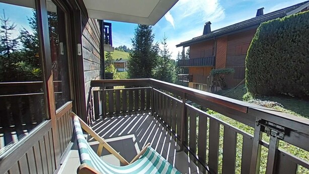 A balcony with a wooden railing, a striped lounge chair, and a view of neighboring houses and greenery. - Cozy 2-room apartment, 6 beds, near slopes (Le Grand-Bornand)