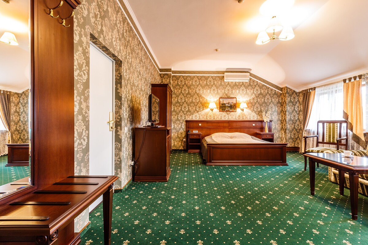 A hotel room with a large bed, a desk, a chair, and a small table. There is a mirror on the wall and a window with curtains.