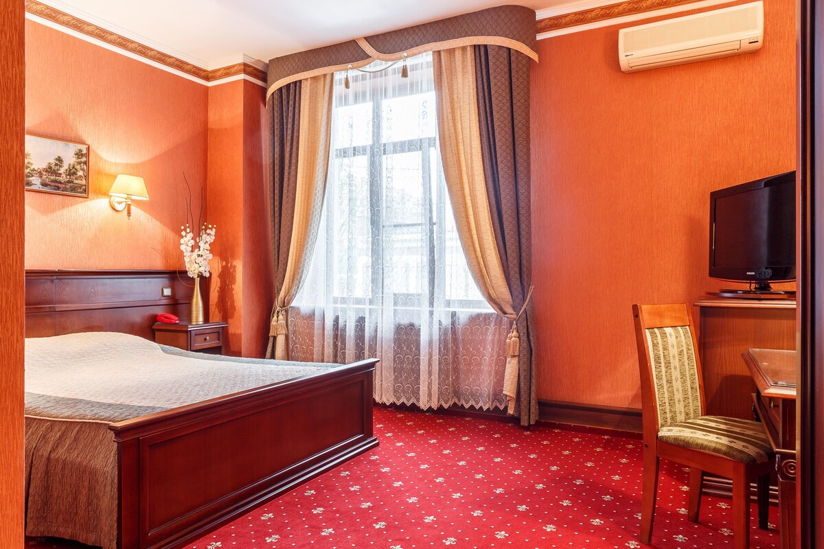 A hotel room with a wooden bed, a desk with a chair, a television, and a window with curtains.