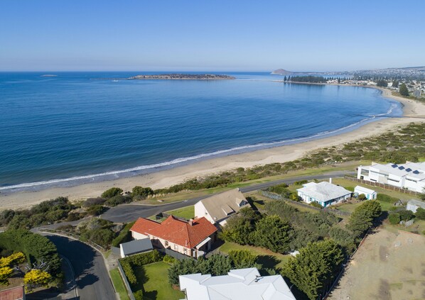 An aerial view of a coastal property with a beach, a road, and a large house with a red roof.