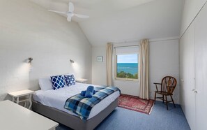 A bedroom with a bed, a chair, a nightstand, a ceiling fan, a window with a view of the sea, and a red rug.