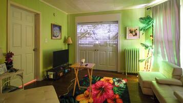 A living room with a television, a small table, a sofa, a floral rug, and a door leading outside.