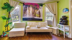A room with a large window, a sofa, a wall-mounted clock, and a decorative waterfall painting.