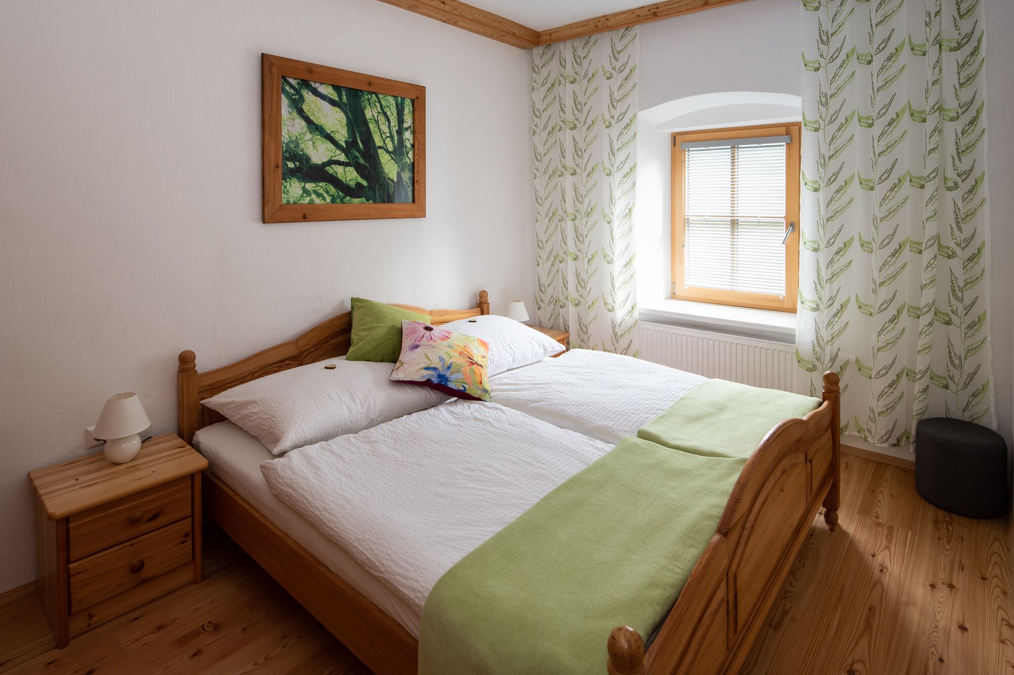 Ein Doppelbett mit Holz Kopfteil, zwei Kissen, eine grüne Tagesdecke und ein Nachttisch mit Lampe. Ein Fenster mit Jalousien und ein Heizkörper sind darunter zu sehen.