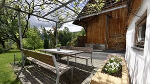 Een houten terras met een bankje en tafel, omringd door groen en een houten gebouw.