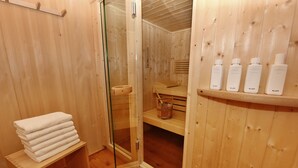 Een houten sauna met een glazen deur, een stapel handdoeken en flessen KLAFS producten op een plank.