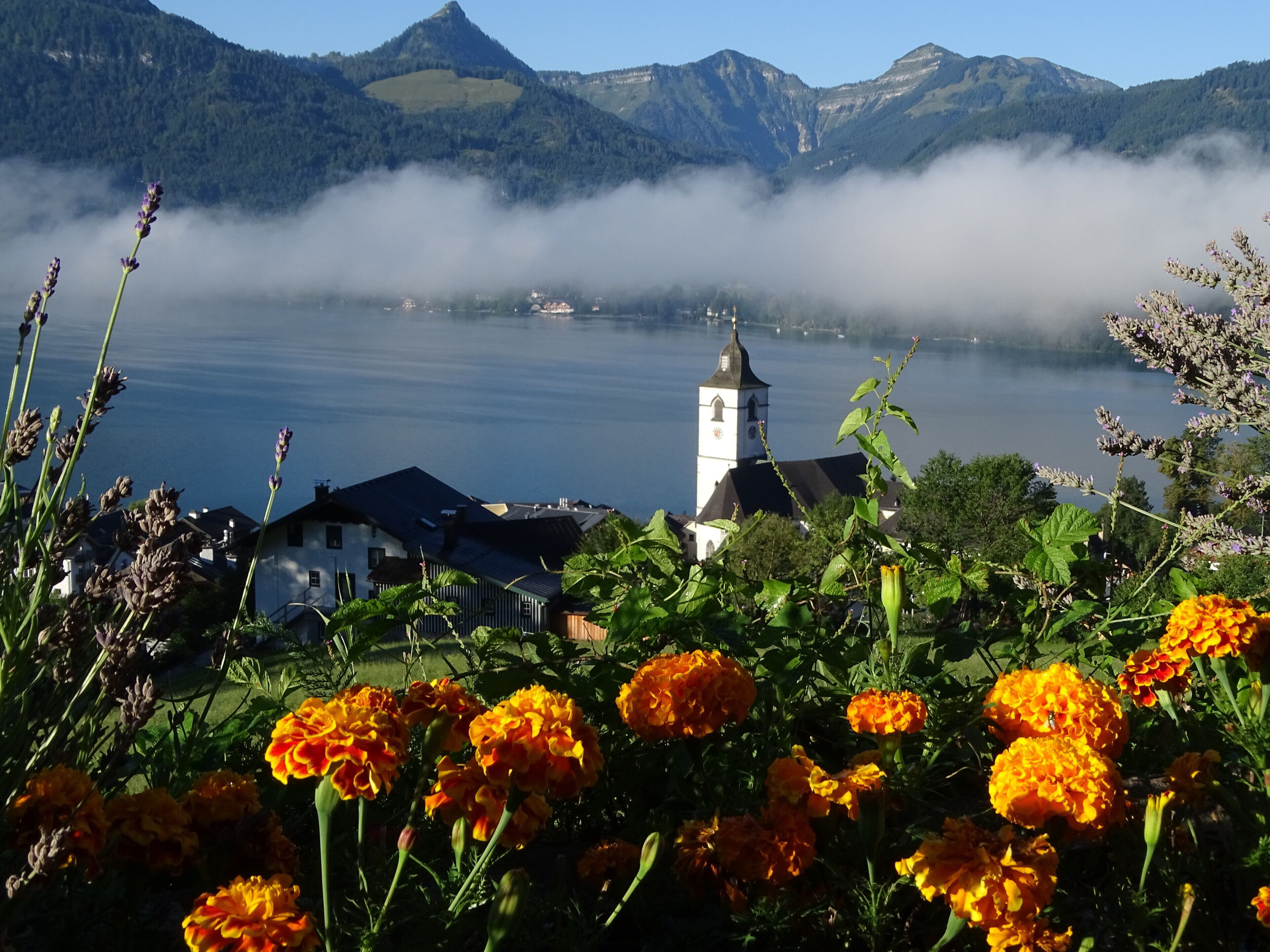 Ein See mit Kirche und Häusern im Hintergrund, umgeben von Bergen und Nebel.